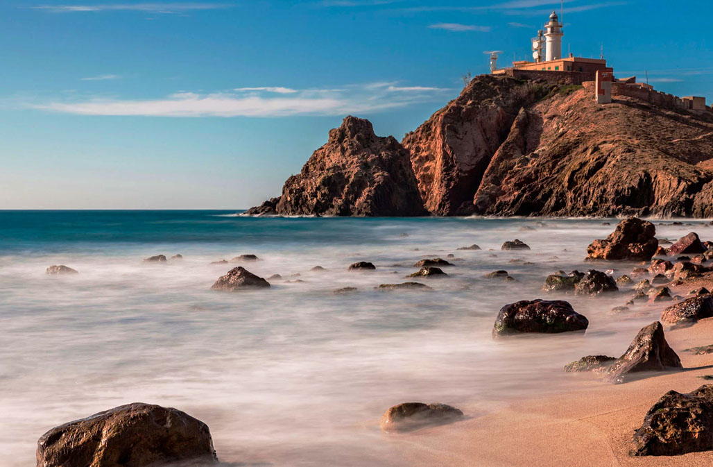 Serenely captured shoreline of Cabo de Gata natural park with a prominent lighthouse perched on rocky cliffs, overlooking misty waves crashing on the scattered boulders.