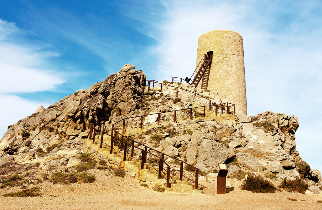 A rustic stone tower perched on a rugged hill at Macenas, with a staircase leading up to the entrance. The sky is clear and blue, and there's a metal railing along the path for safety.