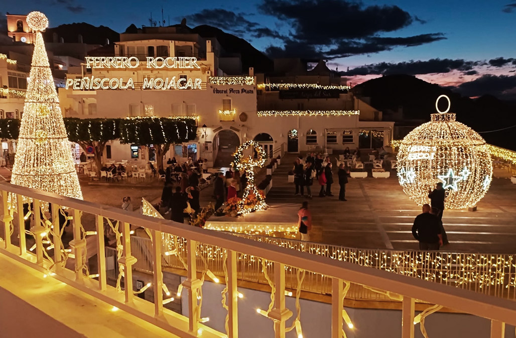 Twilight view of the Mojácar plaza adorned with Ferrero Rocher Christmas decorations, featuring a large, illuminated Christmas bauble and cone-shaped tree made of lights. People are gathered, enjoying the holiday display, with warm golden lighting enhancing the festive atmosphere against a dusky sky.