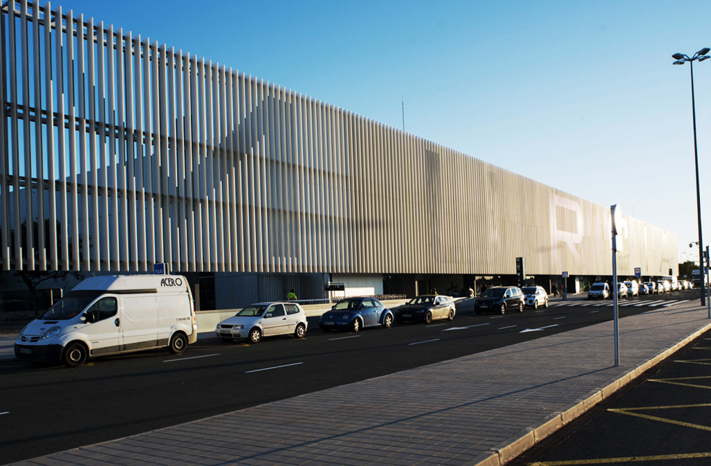 Exterior view of Murcia Airport with a modern, minimalist design featuring a long, horizontal building with vertical metal slats. Cars are parked in front, in the pick-up and drop-off area.