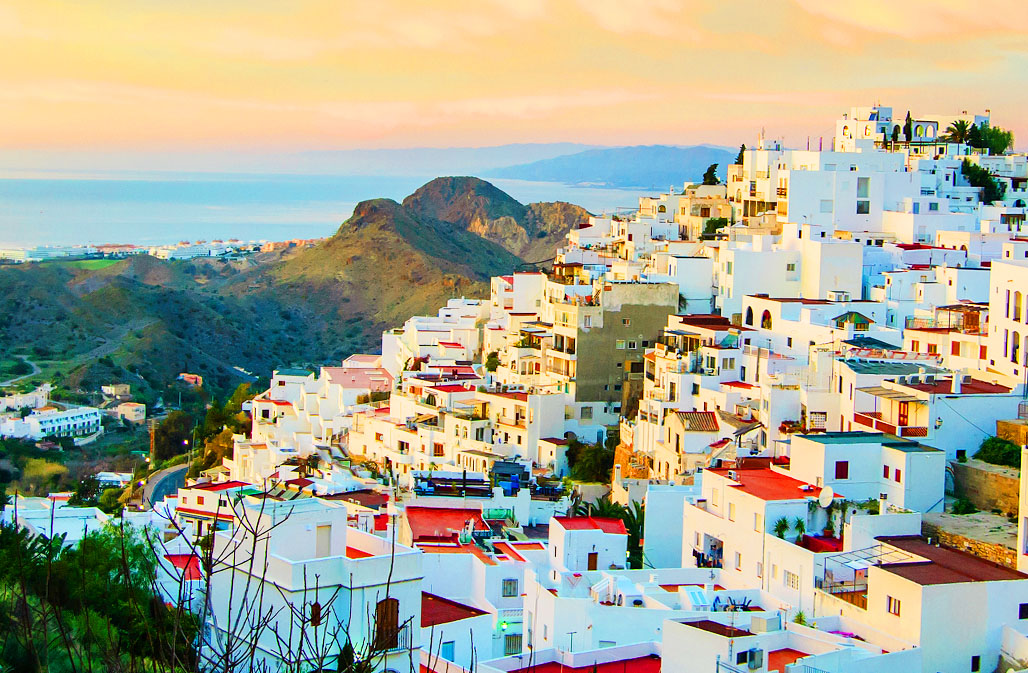 Breathtaking view of a Mojácar Pueblo with multilayered whitewashed buildings, narrow winding streets, and terraces, bathed in the warm glow of sunset, overlooking the serene coastline and the vast sea