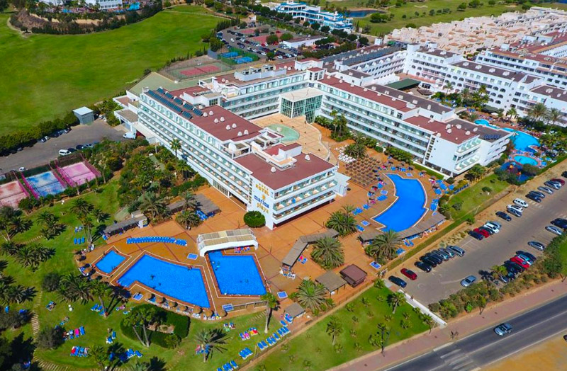 Aerial view of the Marina Hotel complex in Mojácar showing multiple swimming pools surrounded by palm trees, with adjacent golf courses and residential buildings.