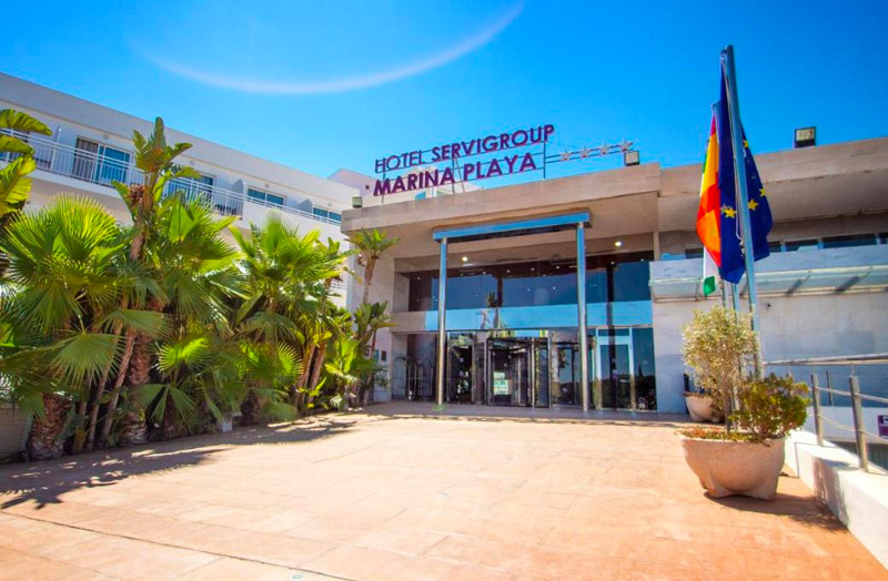 Exterior of Hotel Servigroup Marina Playa with palm trees flanking the main entrance and European Union and Spanish flags on the right.