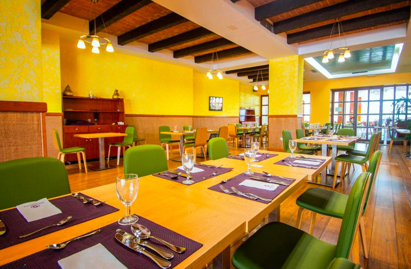 Bright and welcoming dining area at Servigroup Marina Hotel in Mojacar with green chairs, wooden tables set for a meal, and yellow patterned wallpaper, complemented by natural light from large windows and warm overhead lighting.