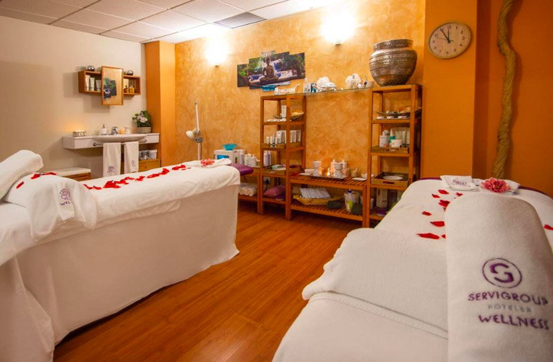 A serene spa treatment room with two white massage tables adorned with red rose petals and towels embroidered with the "Servigroup Hotels Wellness" logo. Shelves stocked with various spa products, a clock on the wall, and a warm orange hue on the walls create a relaxing atmosphere.