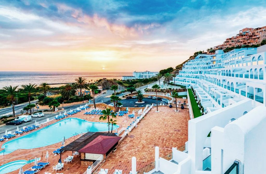 Mojacar Beach at sunset, viewed from a resort balcony, displaying a weather-perfect scene. The horizon is graced by a sun dipping beneath the skyline, casting hues of orange and pink across the sky